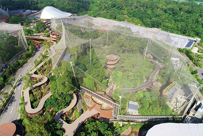 Alt text: “Aerial view of a large cable-supported mesh enclosure surrounded by dense greenery and winding pathways.
