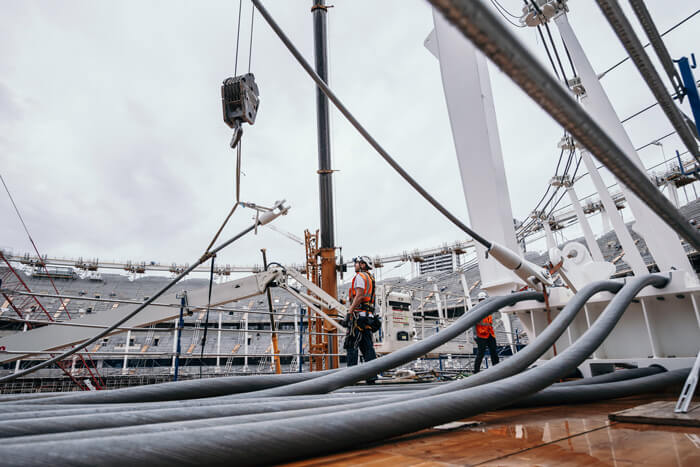 Construction workers installing large structural cables at a stadium using a crane.