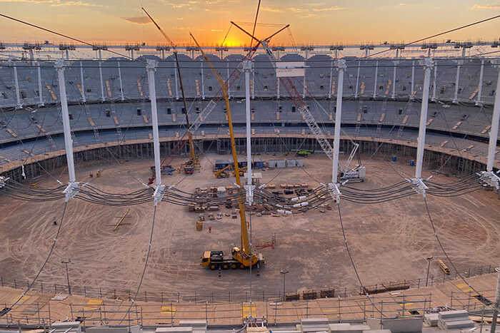 Construction of a large stadium roof with cranes installing a cable-supported tensile structure at sunset.