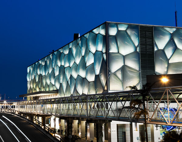 Illuminated building with a geometric translucent facade beside an elevated walkway at night