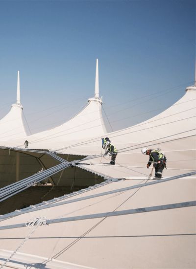 Workers wearing safety harnesses installing and maintaining a large tensile membrane roof structure.